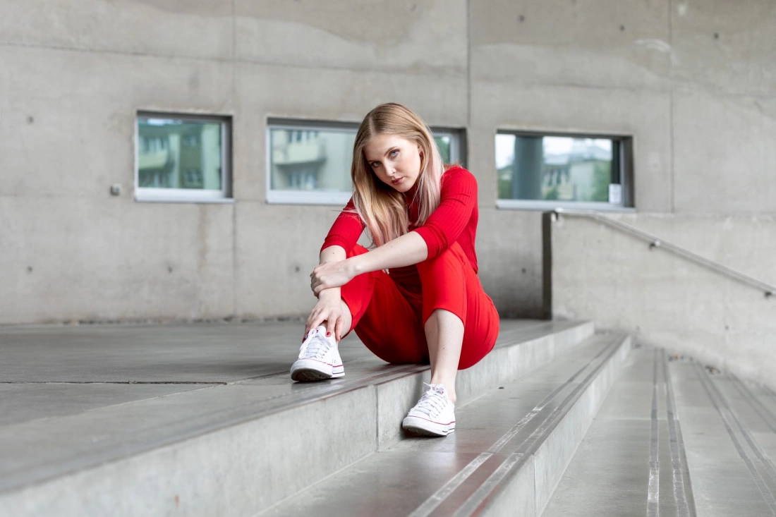 Kobieta w czerwonym stroju siedzi na schodach w minimalistycznej przestrzeni.