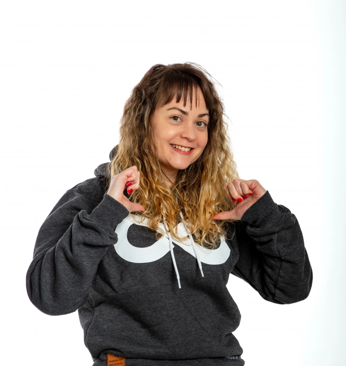 Kobieta z falującymi włosami w szarej bluzie uśmiecha się i wskazuje na siebie.