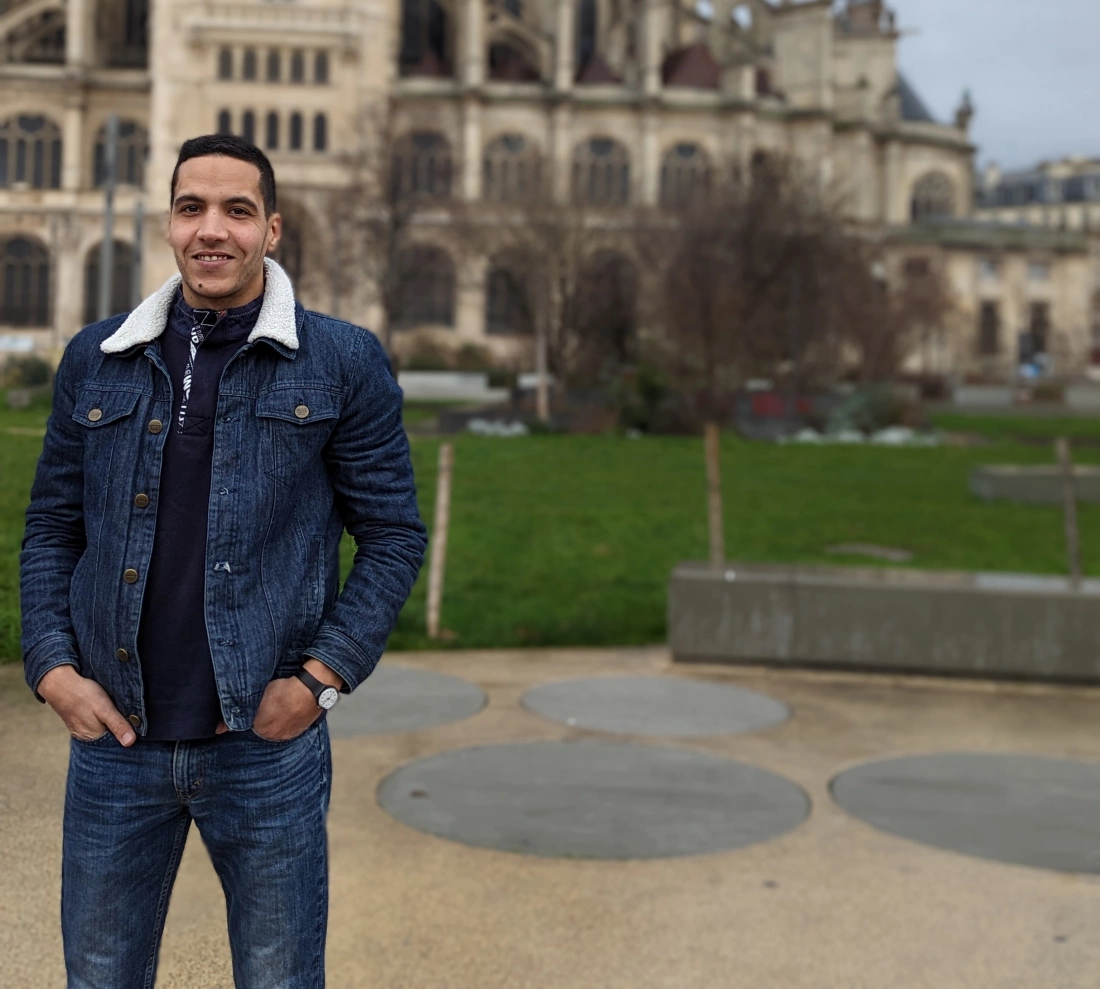 Mężczyzna w dżinsowej kurtce i niebieskich dżinsach stoi w parku przed budynkiem.