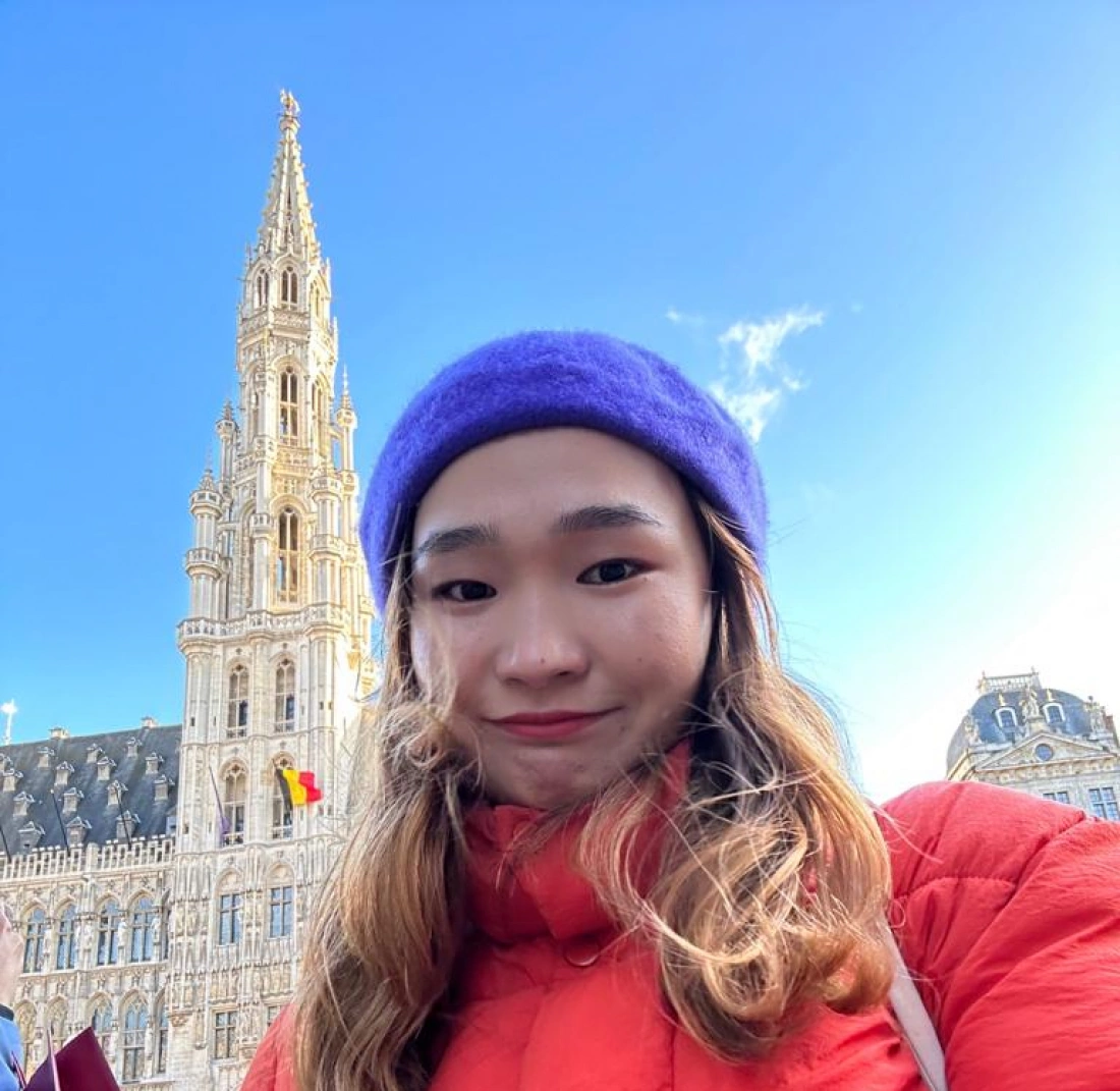 Osoba w kolorowej kurtce i czapce stoi przed dużym, zabytkowym budynkiem na tle nieba.