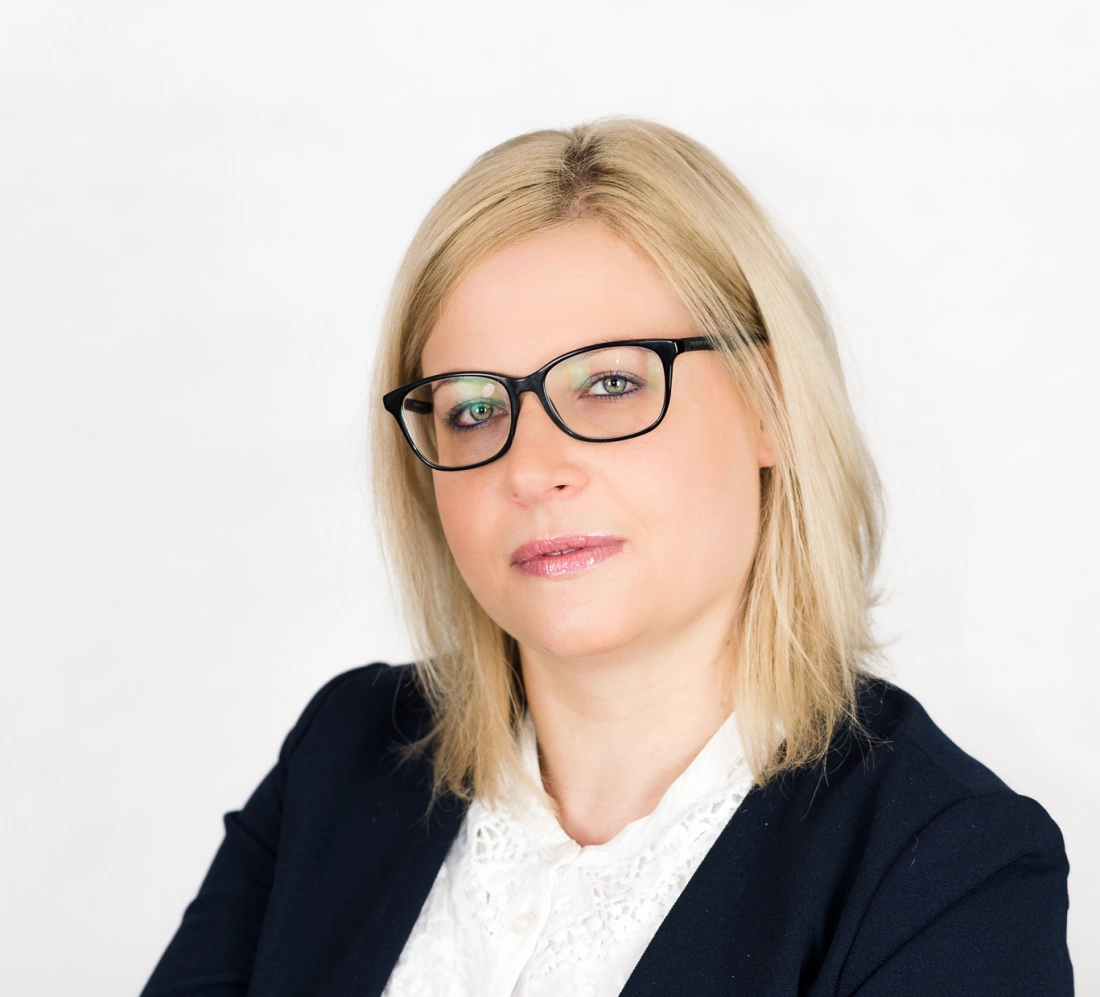 Osoba z jasnymi włosami, w okularach, uśmiechająca się w niewielkim skupieniu. W tle jasna ściana.