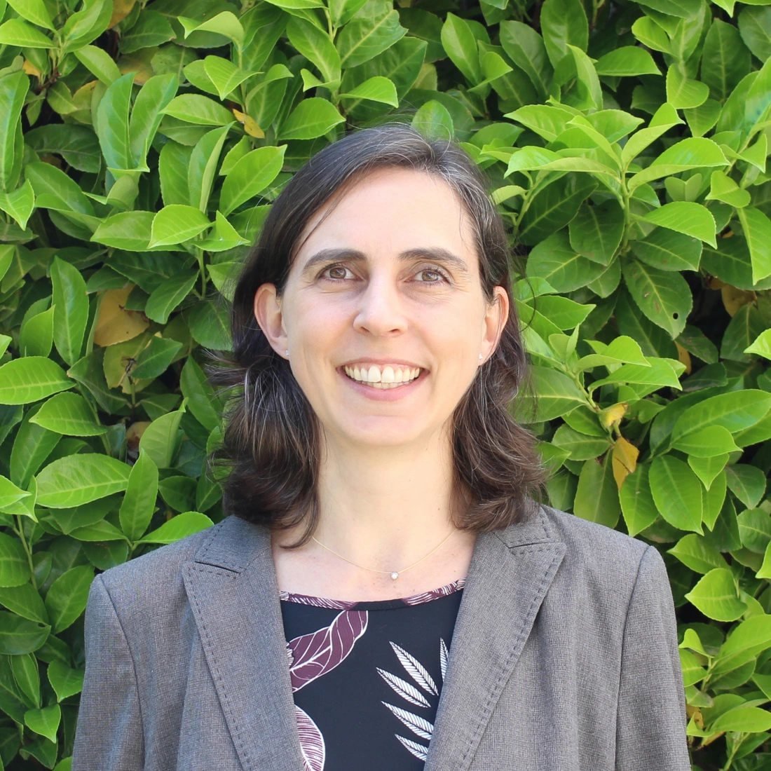 Kobieta w szarej marynarce uśmiecha się na tle zielonych liści.