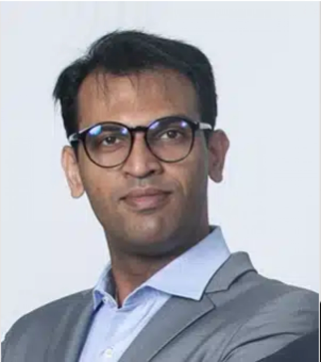 Mężczyzna w okularach, ubrany w szary garnitur, z neutralnym wyrazem twarzy.