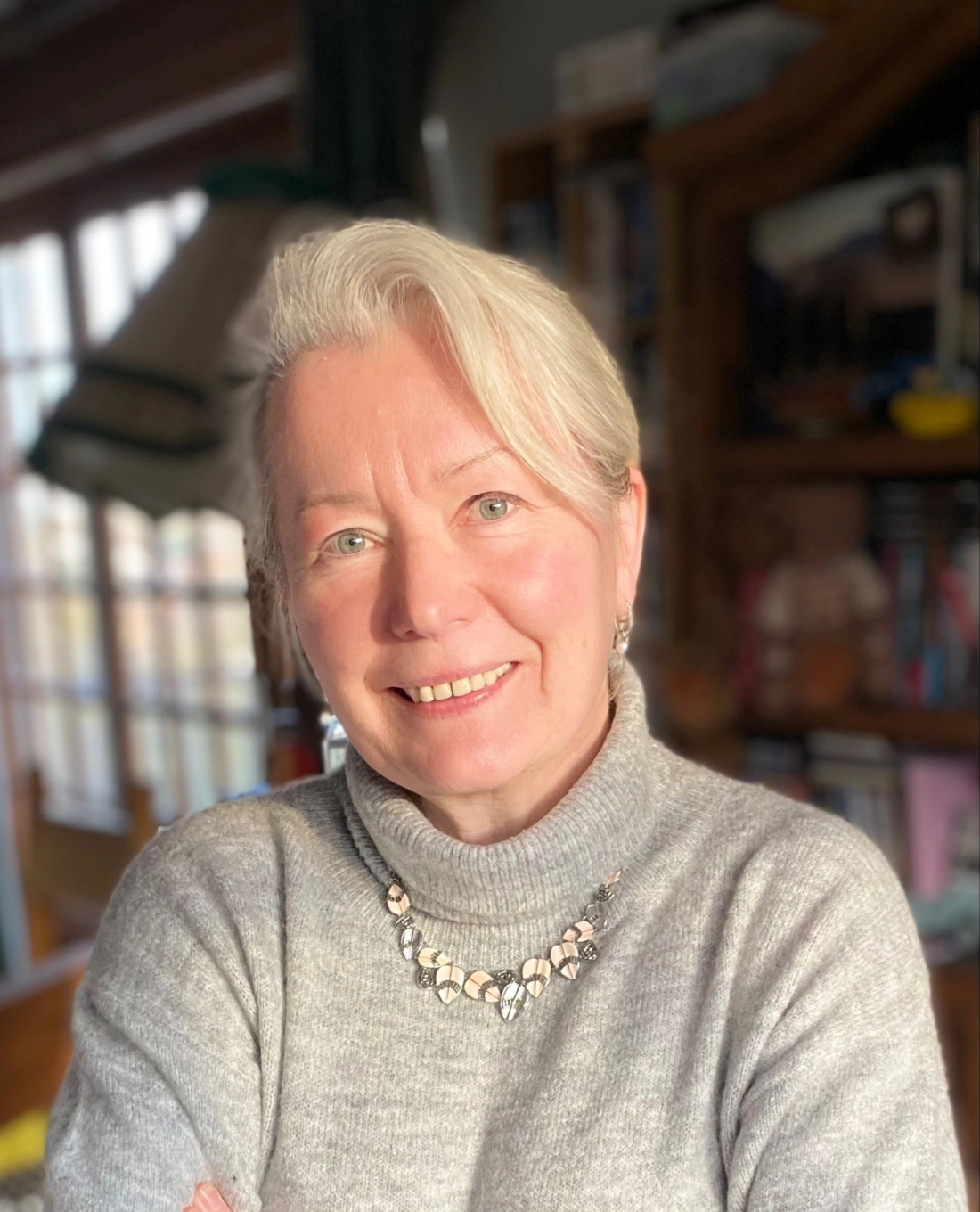 Uśmiechnięta kobieta w szarym swetrze z naszyjnikiem, stojąca w jasnym pomieszczeniu.