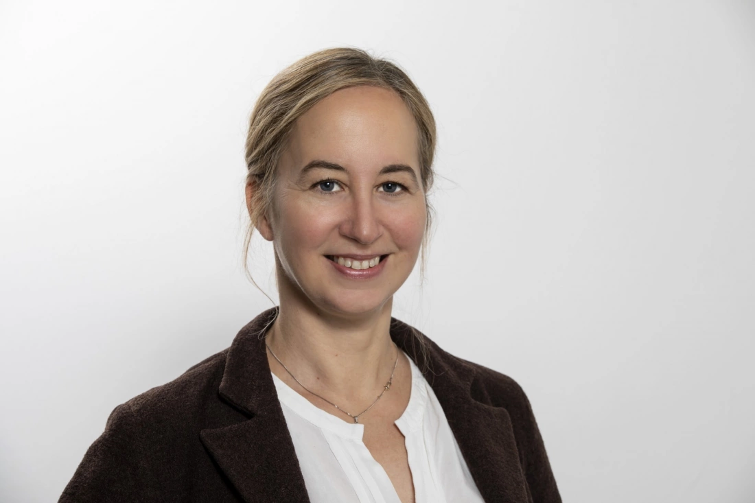 Kobieta uśmiechająca się, ubrana w brązowy płaszcz, na tle jasnej ściany.