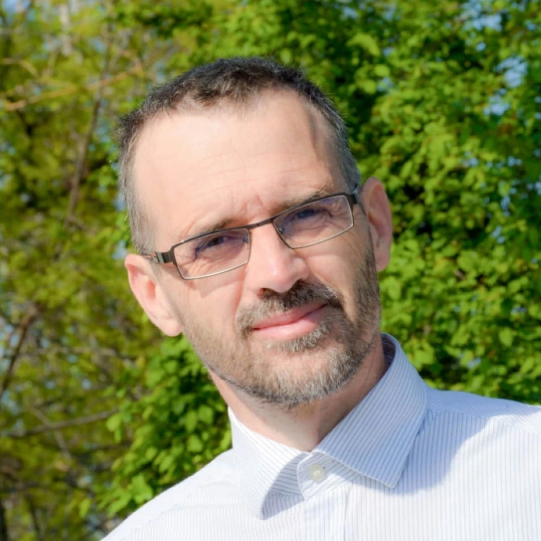 Mężczyzna z okularami, uśmiechający się na tle zielonych drzew.