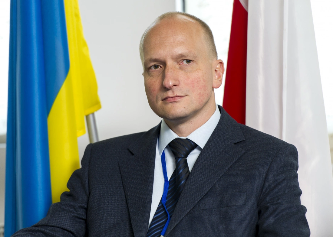 Mężczyzna w garniturze, siedzący w biurze, z flagami Ukrainy i Polski w tle.