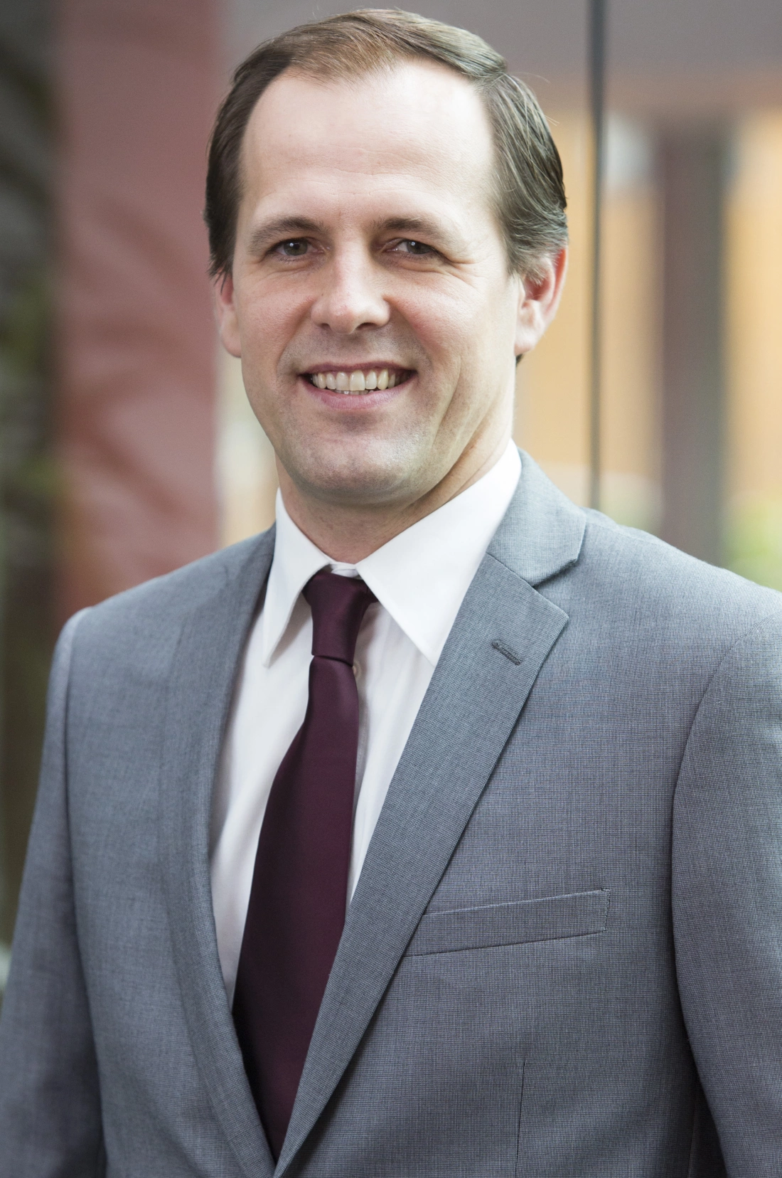 Mężczyzna w szarym garniturze i bordowej krawacie, uśmiechający się w biurowym otoczeniu.