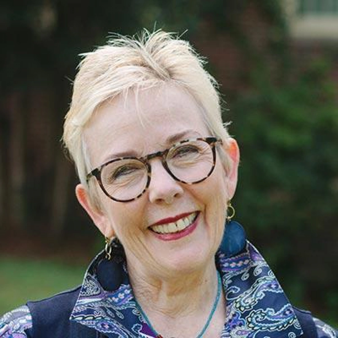 Uśmiechnięta kobieta w okularach, z krótkimi blond włosami, nosząca kolorowy strój.