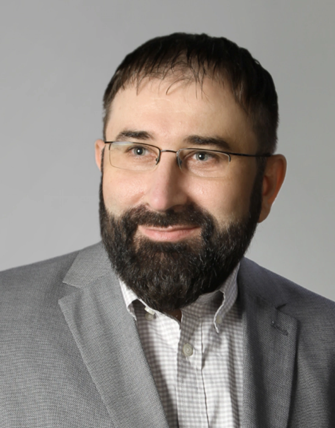 Mężczyzna z brodą, w okularach i szarym garniturze, uśmiecha się na tle jednolitego tła.