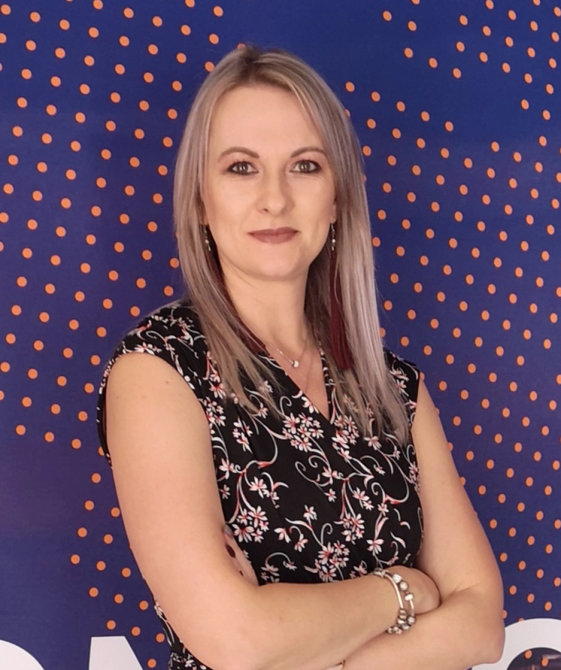 Kobieta z długimi, prostymi włosami i uśmiechem, ubrana w czarną sukienkę w kwiaty, stoi z założonymi rękami przed niebieskim tłem w kropki.