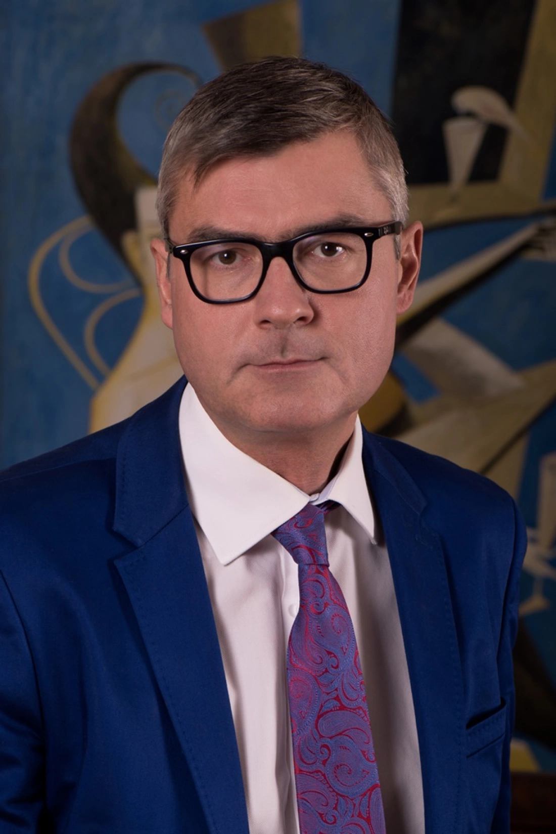 Mężczyzna w niebieskim garniturze i okularach, stojący przed abstrakcyjnym tłem.