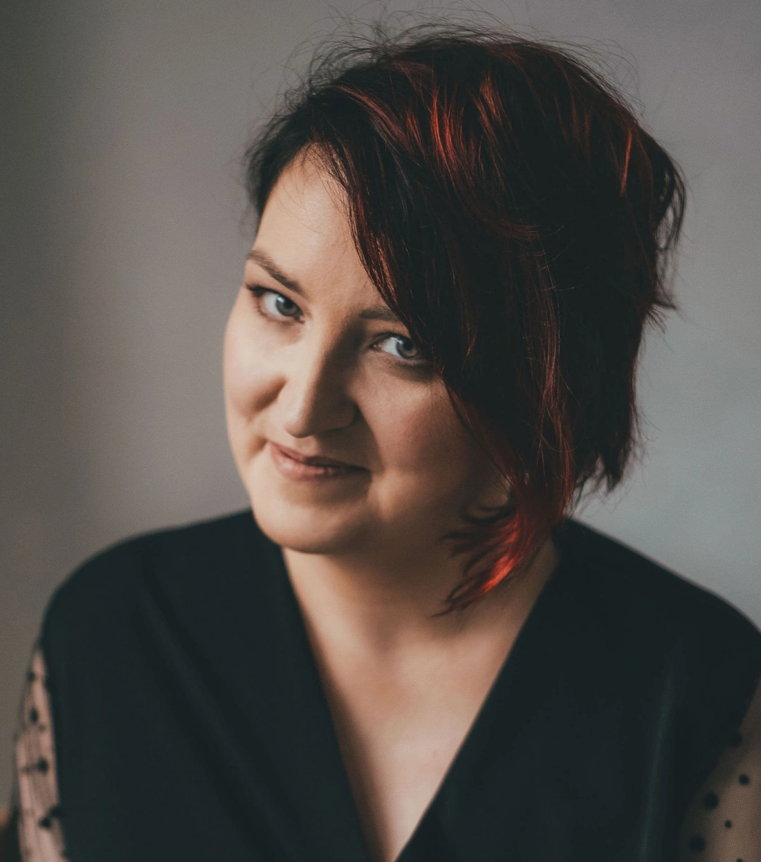 Portret osoby z krótkimi, ciemnymi włosami z czerwonym akcentem, uśmiechającej się w sposób tajemniczy.