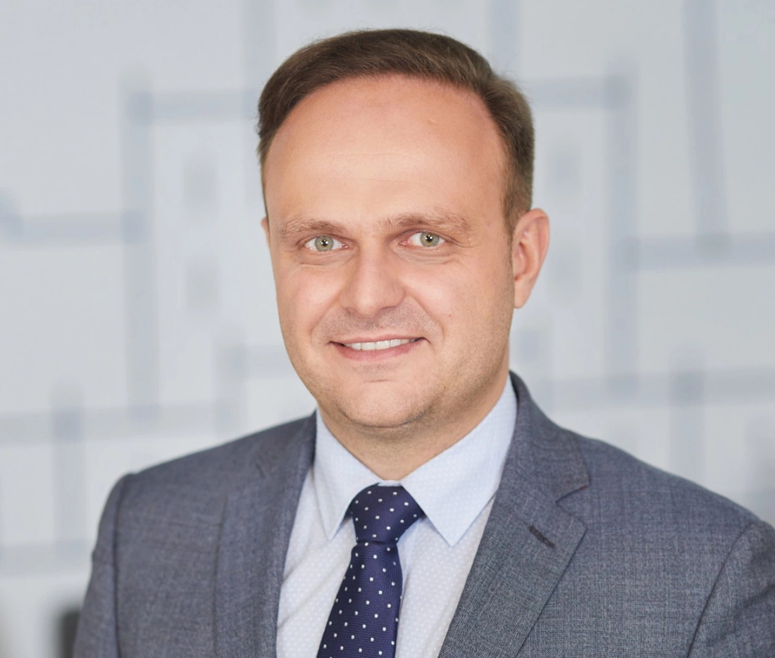 Mężczyzna w garniturze z uśmiechem, stojący przed jasnym tłem.