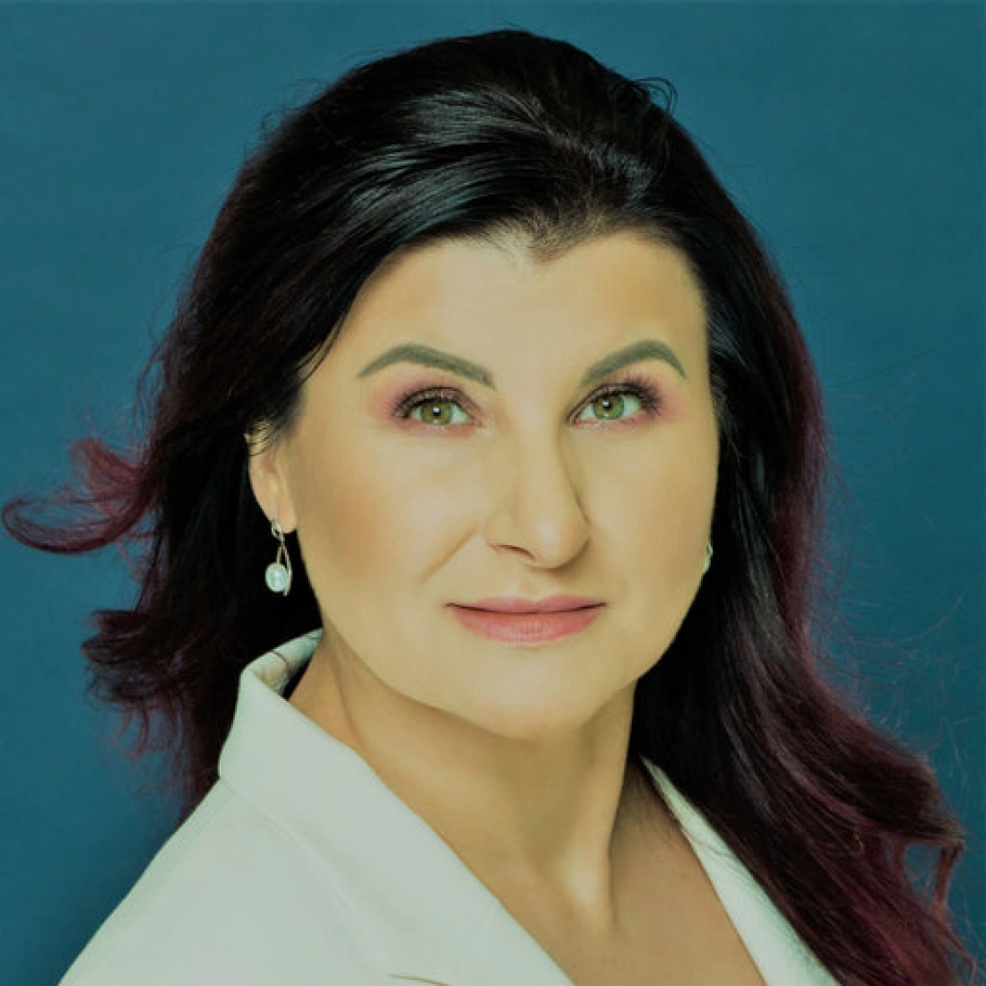 Portret kobiety z długimi, ciemnymi włosami, w białej bluzce, z delikatnym makijażem i zielonymi oczami, na tle w jednolitym kolorze.
