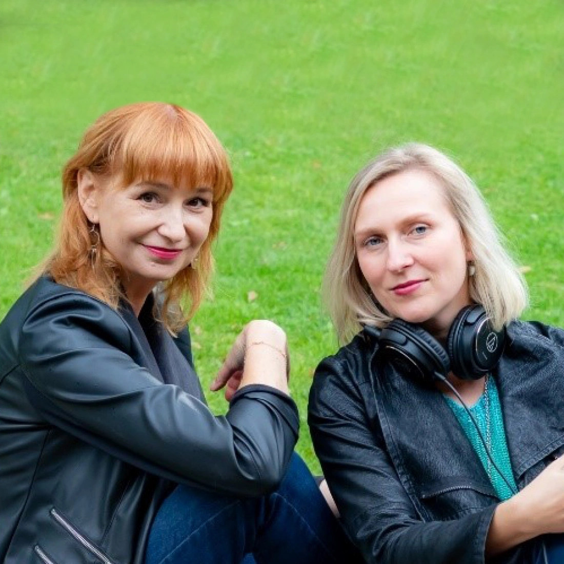 Dwie kobiety siedzą na trawie, uśmiechając się do aparatu.