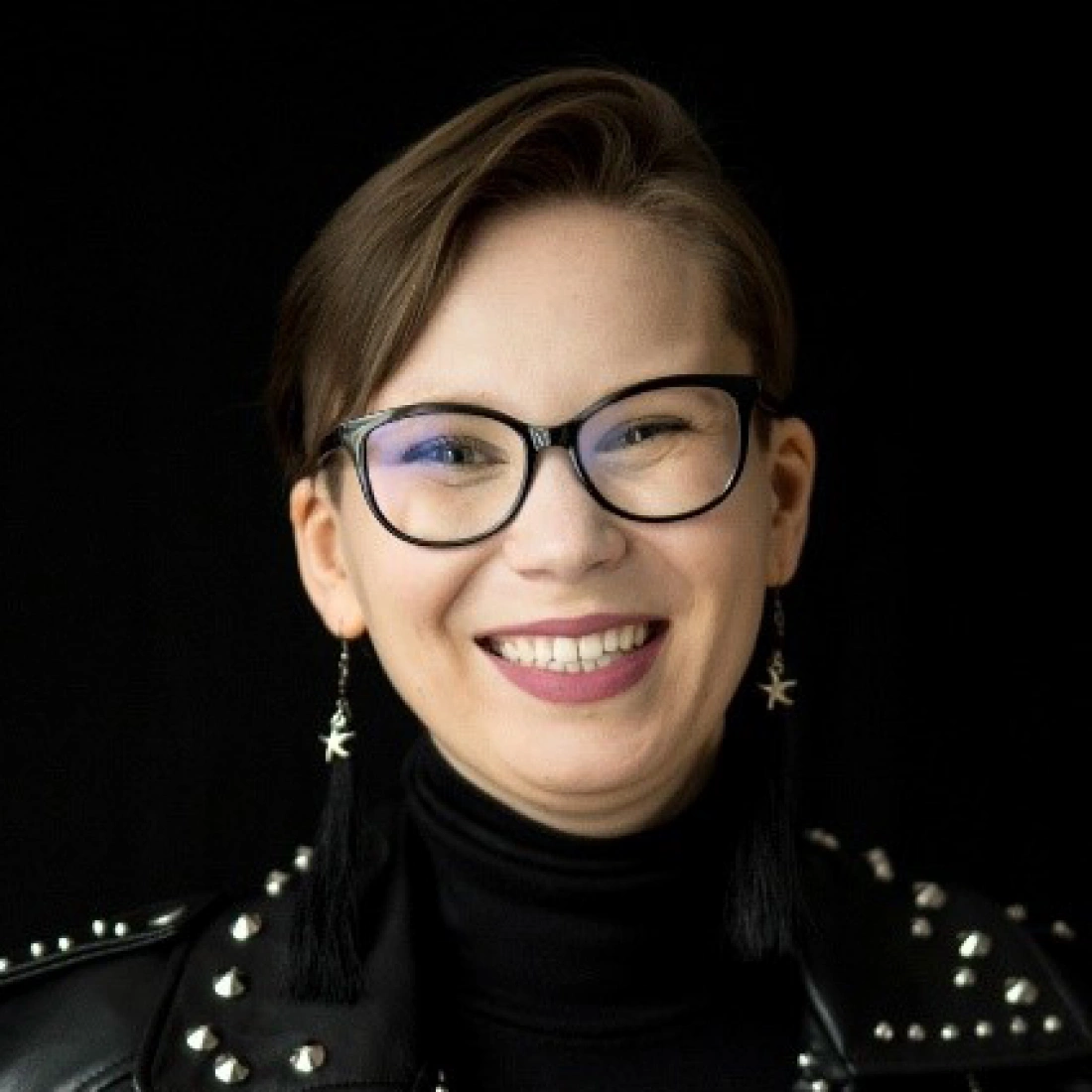 Uśmiechnięta osoba w okularach, nosząca skórzaną kurtkę i długie kolczyki.