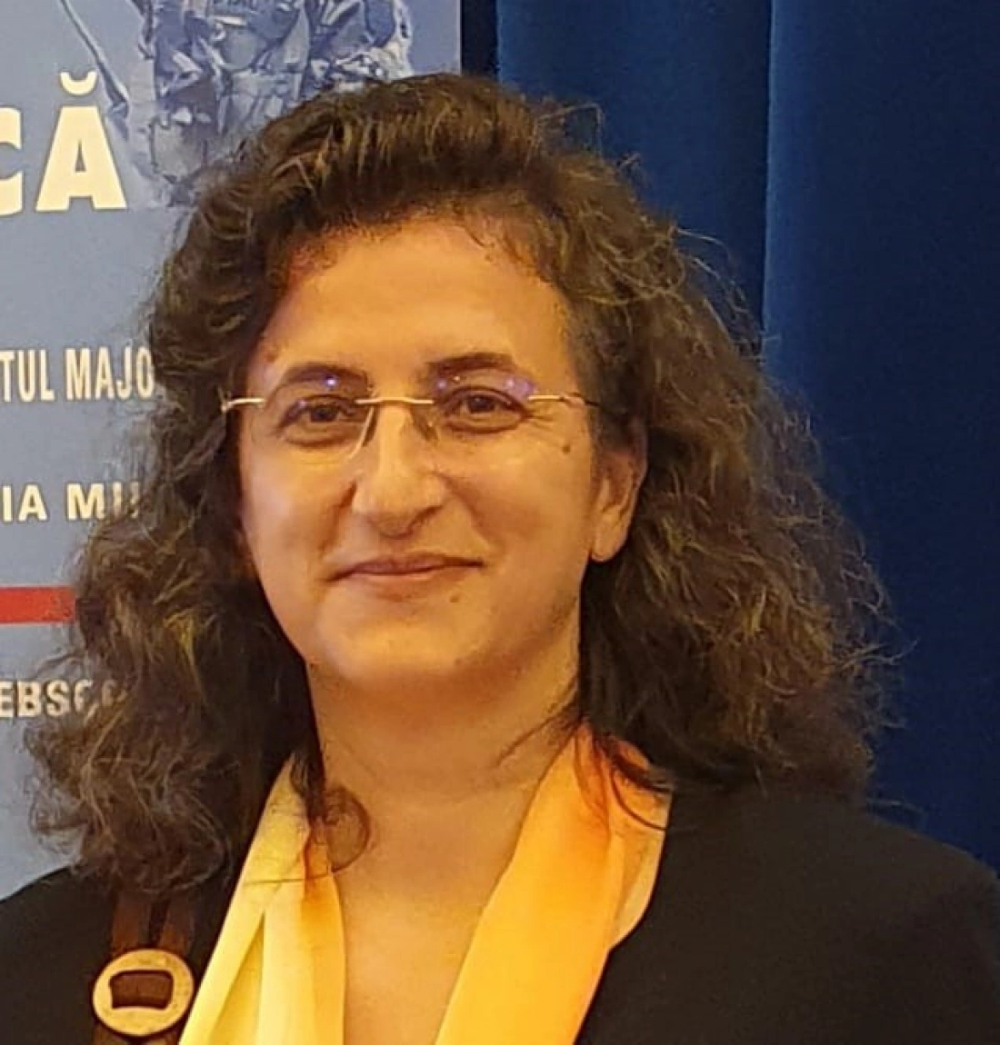 Kobieta z kręconymi włosami, uśmiechająca się, w okularach, na tle niebieskiego tła z plakatem.