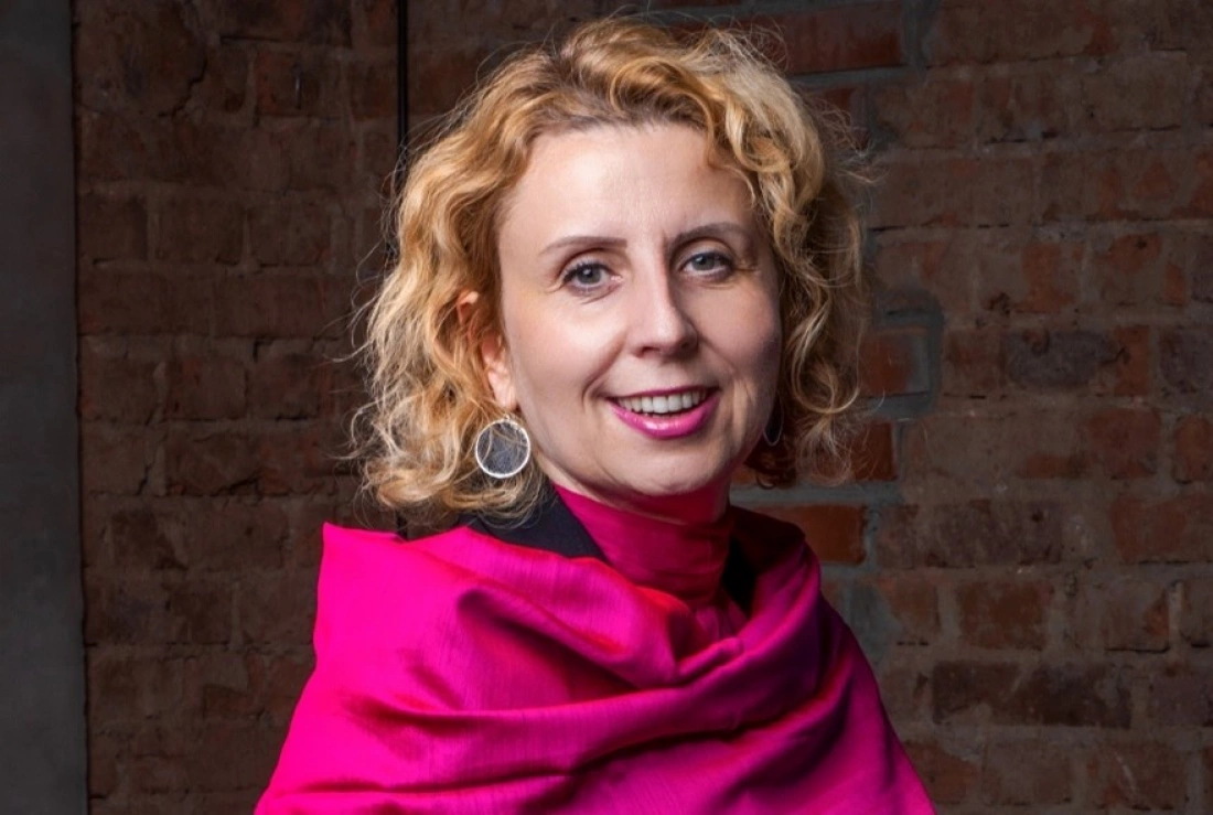 Kobieta z kręconymi włosami, uśmiechająca się w różowym szalu, na tle ceglanej ściany.