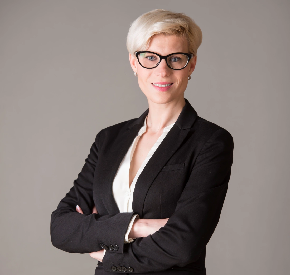 Kobieta w eleganckim garniturze, z krótkimi włosami i okularami, stojąca z założonymi rękami.