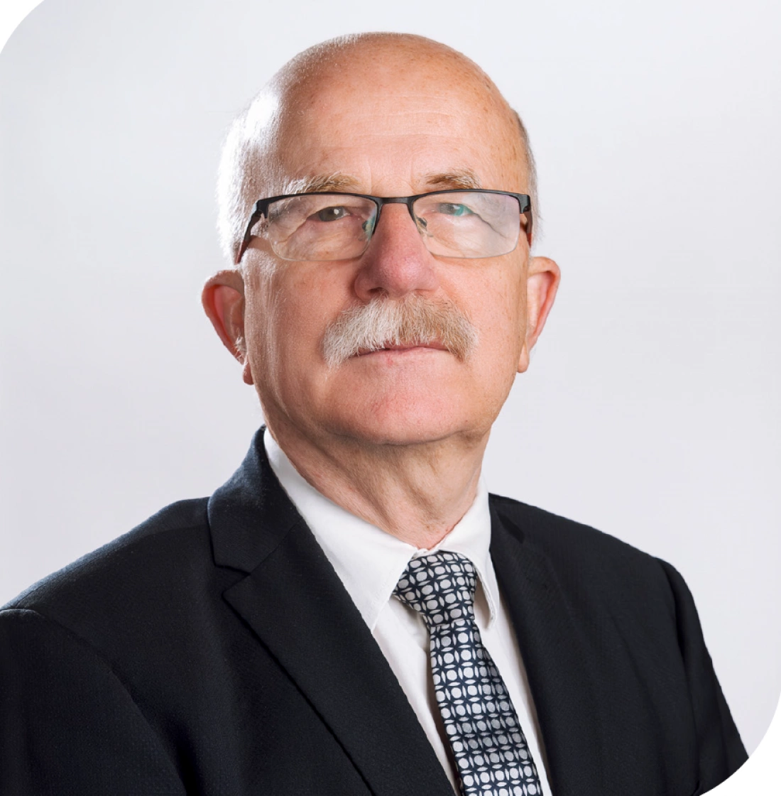 Mężczyzna w okularach, z siwymi włosami i wąsami, ubrany w garnitur i krawat, na tle jasnej, neutralnej ściany.