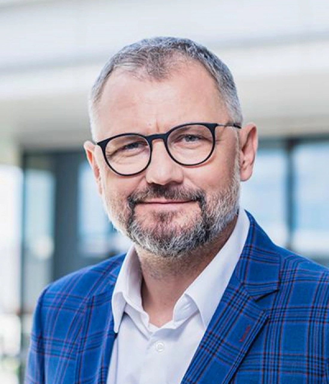 Mężczyzna w okularach, z brodą, w niebieskiej marynarce w kratę, uśmiecha się w nowoczesnym wnętrzu.