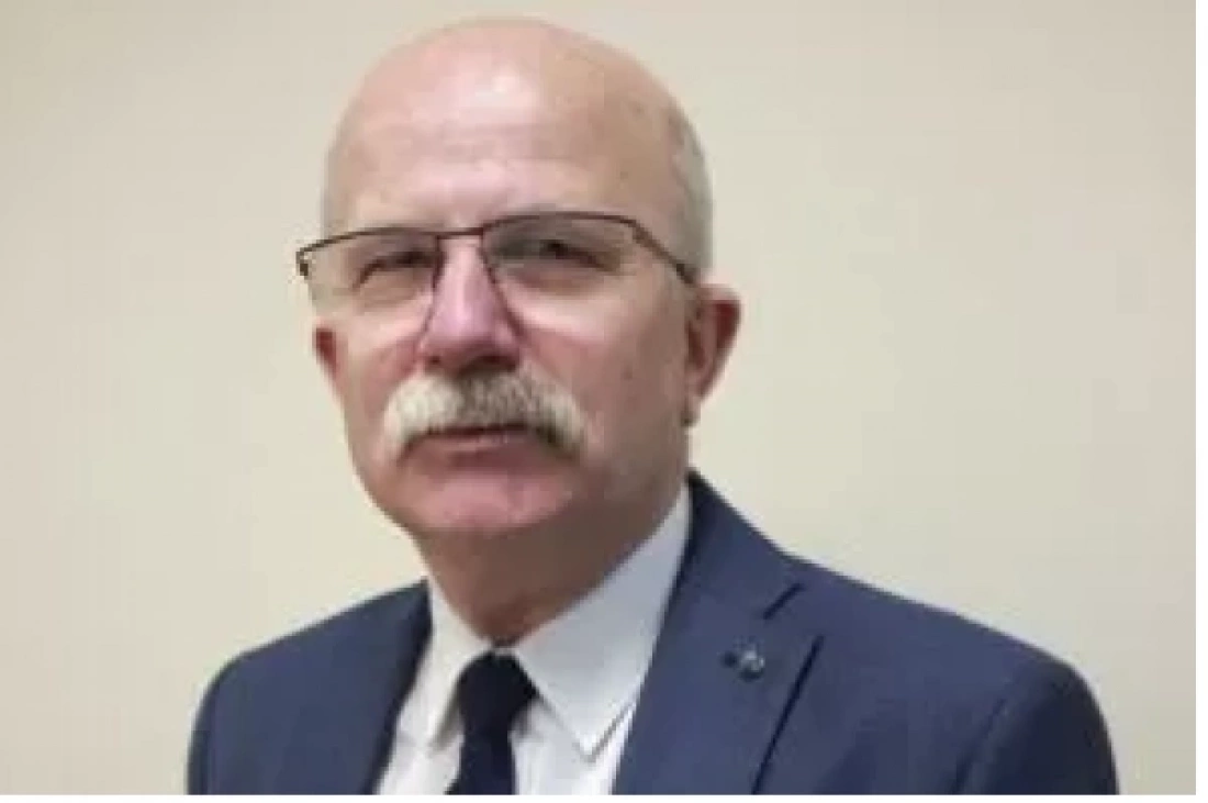 Mężczyzna w okularach, z wąsem, ubrany w granatowy garnitur i krawat, na tle jasnej ściany.