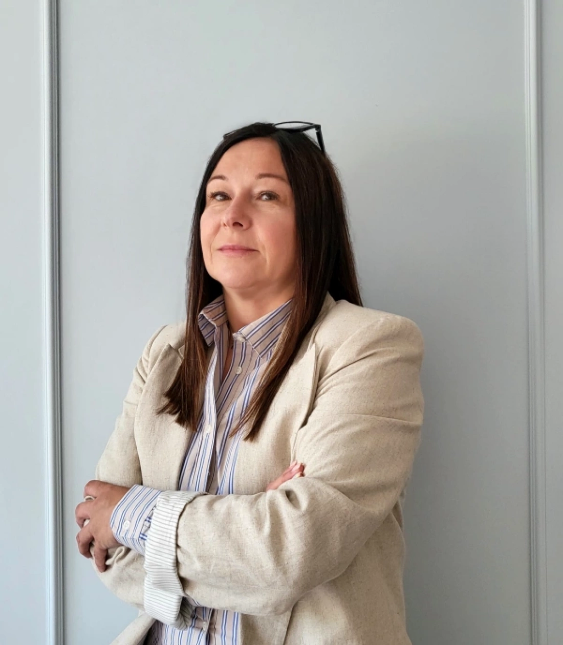 Kobieta w jasnym garniturze z rękami skrzyżowanymi na piersi, stojąca przed szarym tłem.