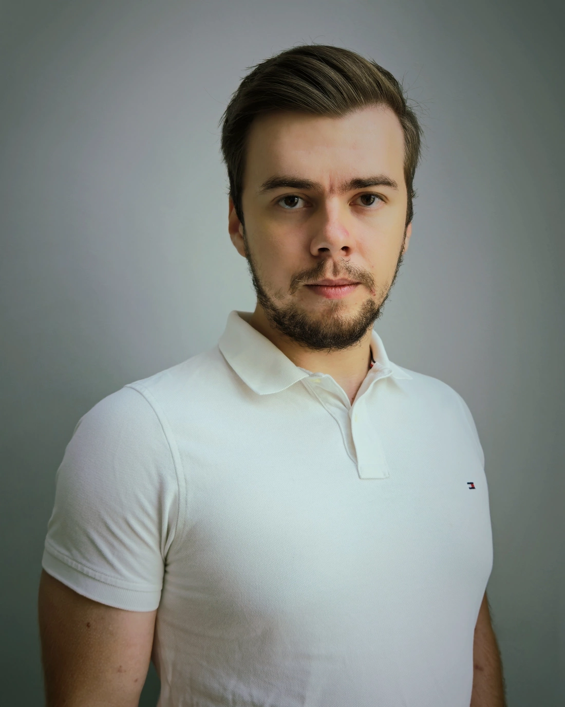 Mężczyzna w białej koszulce polo, z krótkimi włosami i zarostem, patrzy w obiektyw. Tło w stonowanych kolorach.