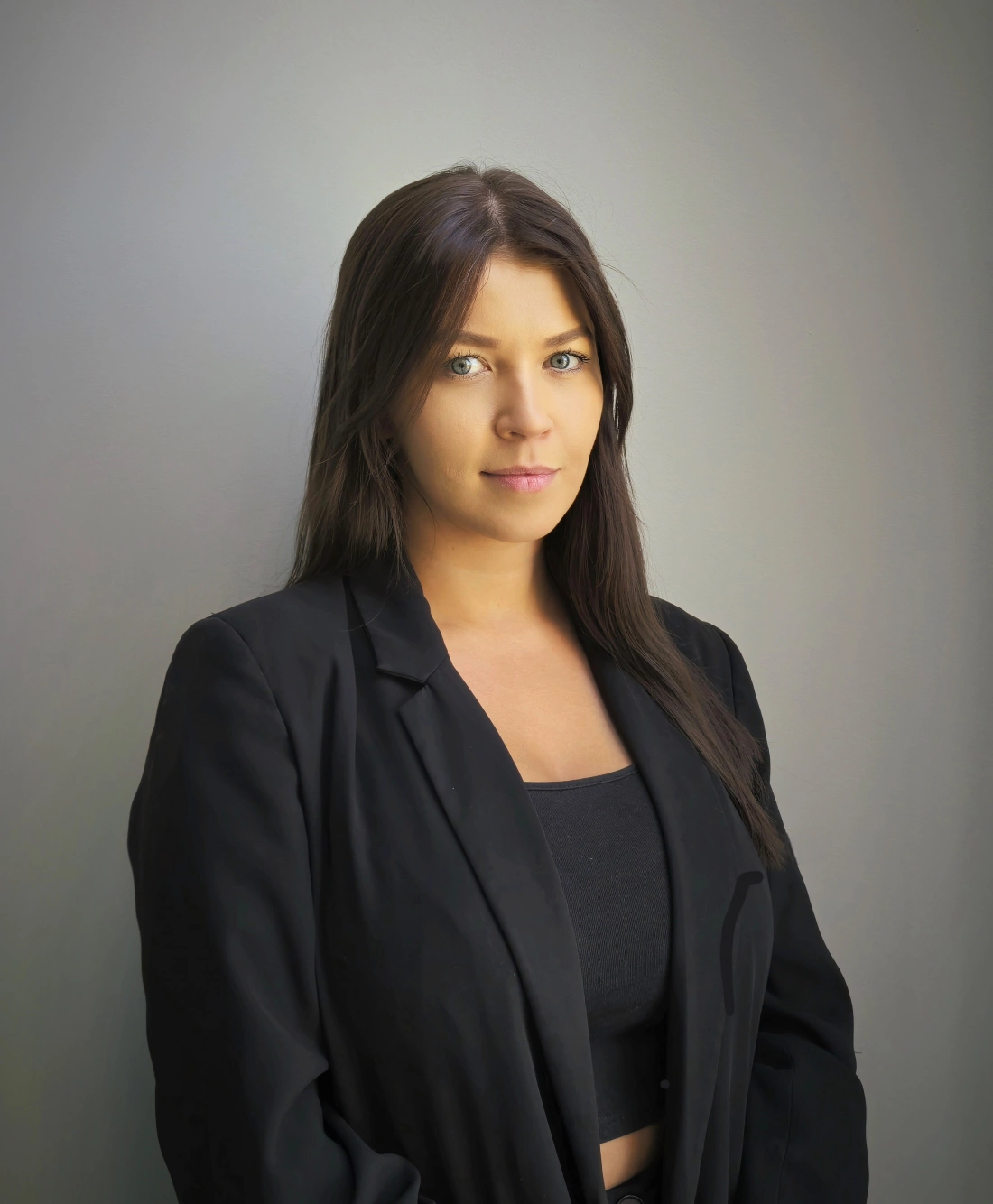 Kobieta w czarnym garniturze z długimi włosami, stojąca na tle szarej ściany.