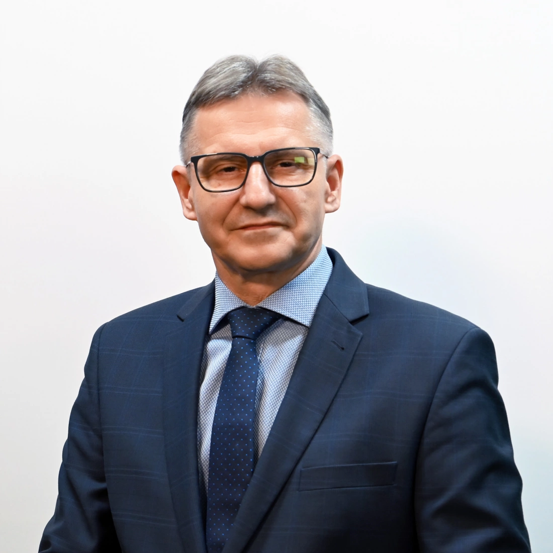 Mężczyzna w eleganckim garniturze, noszący okulary, na tle jasnej ściany.