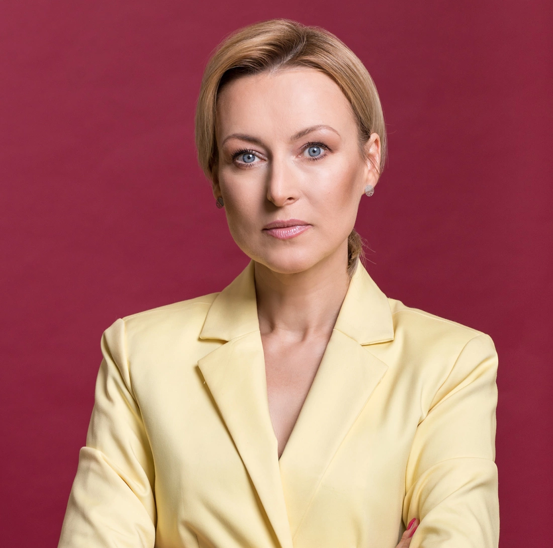 Kobieta w żółtej marynarce, z poważnym wyrazem twarzy, na tle czerwonym.