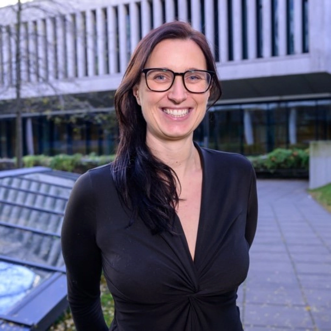 Uśmiechnięta kobieta w czarnej bluzce, stojąca na tle nowoczesnego budynku.