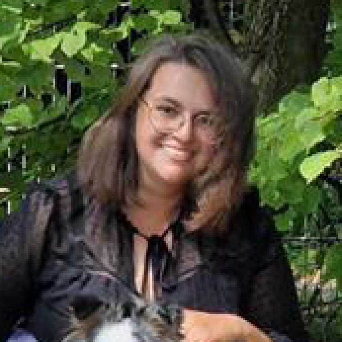 Kobieta w czarnym stroju, uśmiechająca się i trzymająca psa, w otoczeniu zieleni.