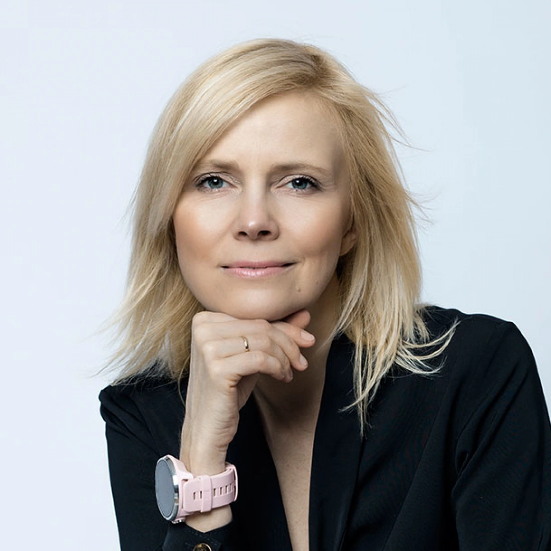 Kobieta z blond włosami w czarnej bluzce, ubrana w różowy zegarek, uśmiecha się, opierając głowę na dłoni.