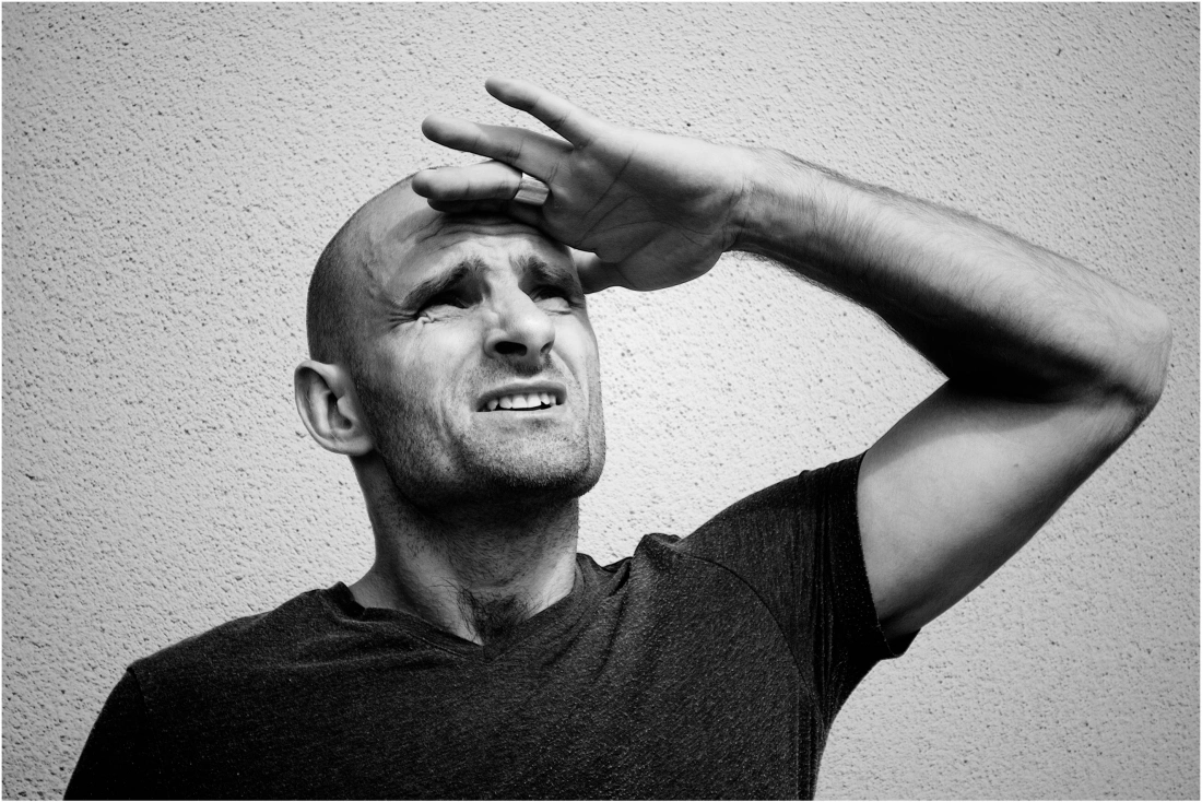 Mężczyzna w szarej koszulce, zasłaniający oczy ręką, patrzy w górę.