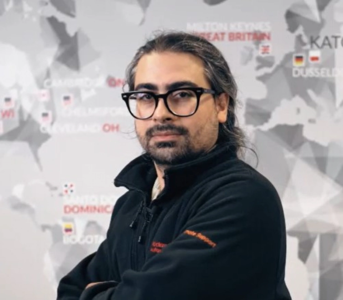 Mężczyzna z długimi włosami i okularami, ubrany w czarną bluzę, stoi przed mapą świata.