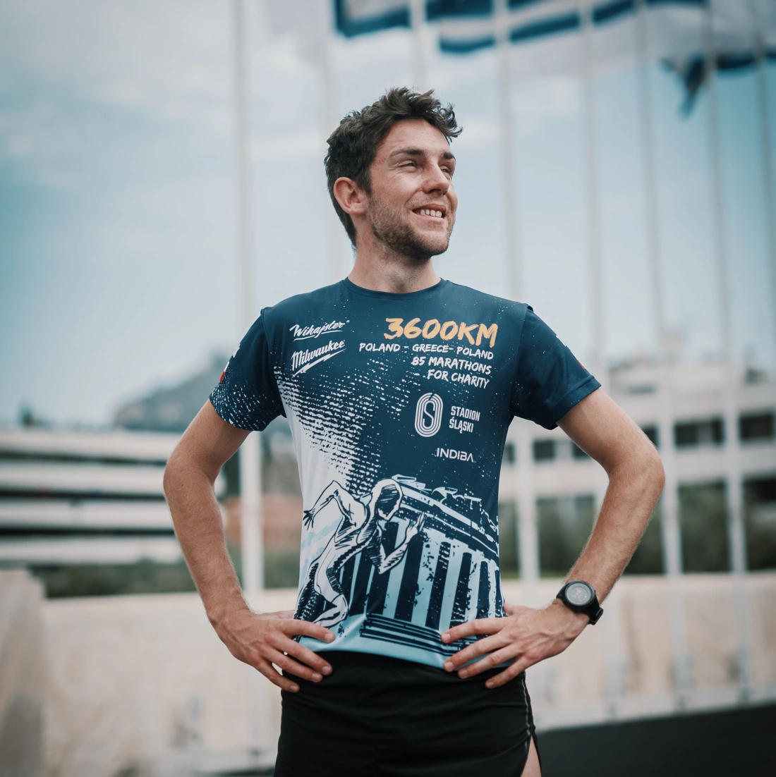 Mężczyzna w sportowej koszulce z grafiką uśmiecha się, stojąc na tle flag.