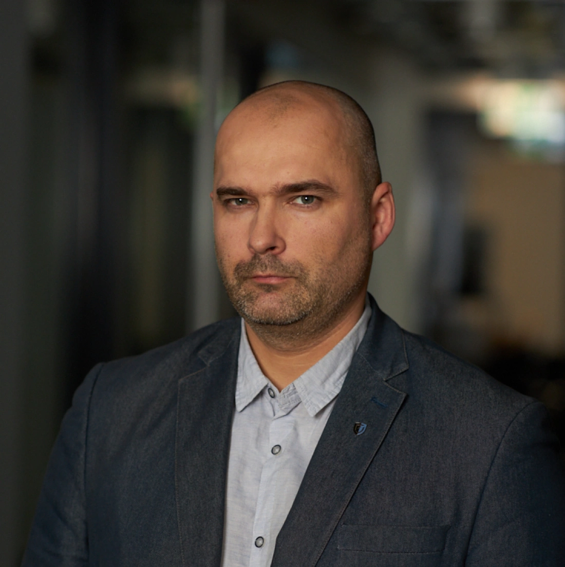 Mężczyzna w garniturze z poważnym wyrazem twarzy, stojący w pomieszczeniu z rozmytym tłem.