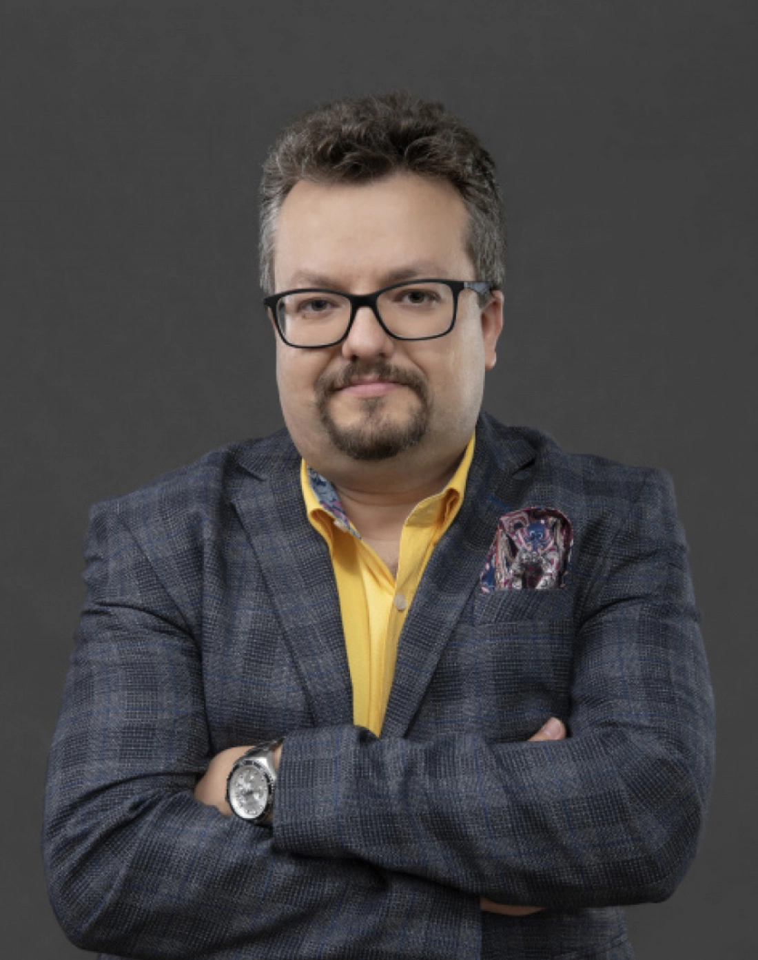 Mężczyzna w okularach, ubrany w granatowy garnitur z kieszonkową chustą, z założonymi rękami.