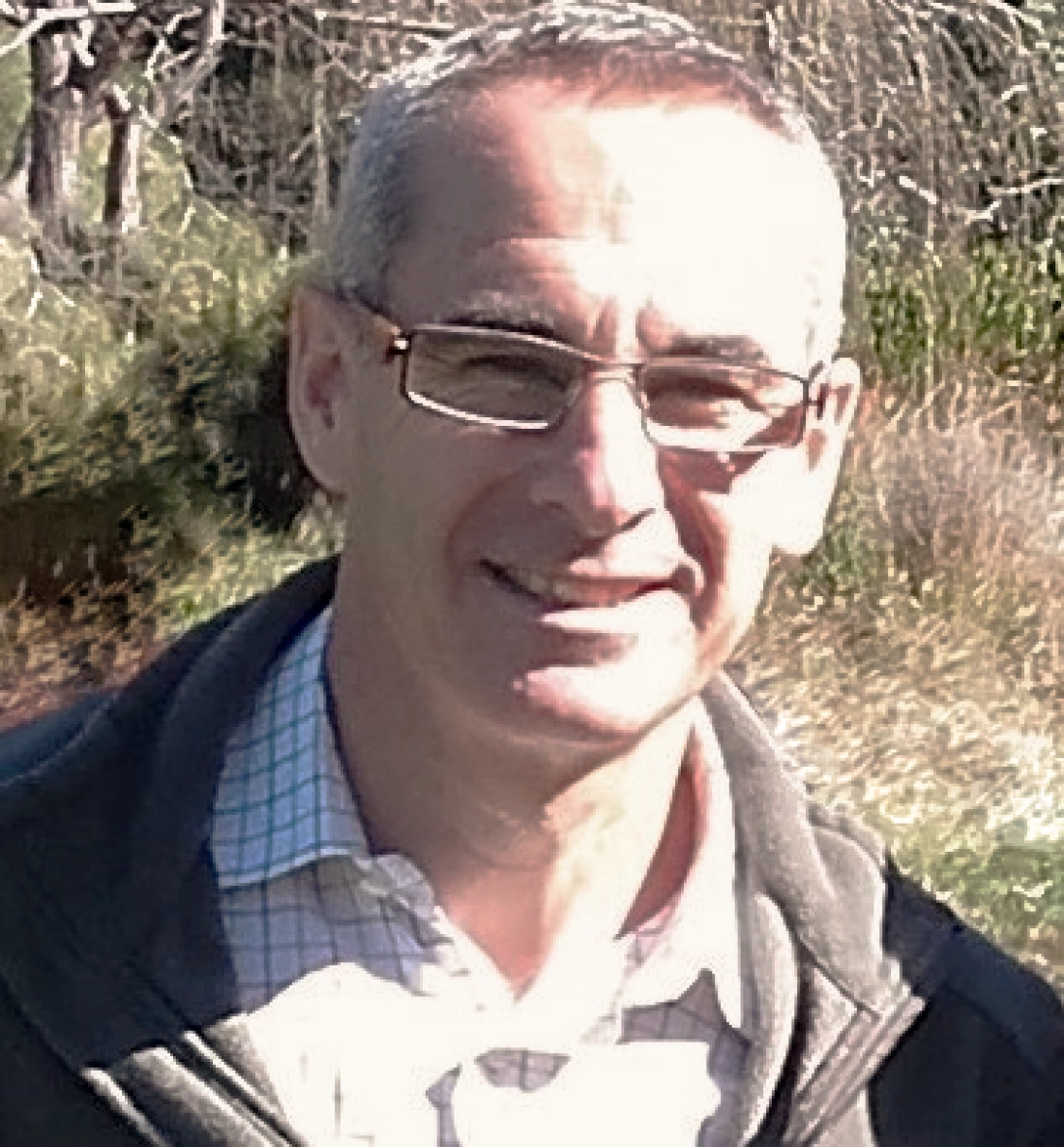 Mężczyzna w okularach uśmiecha się na tle zielonego krajobrazu.