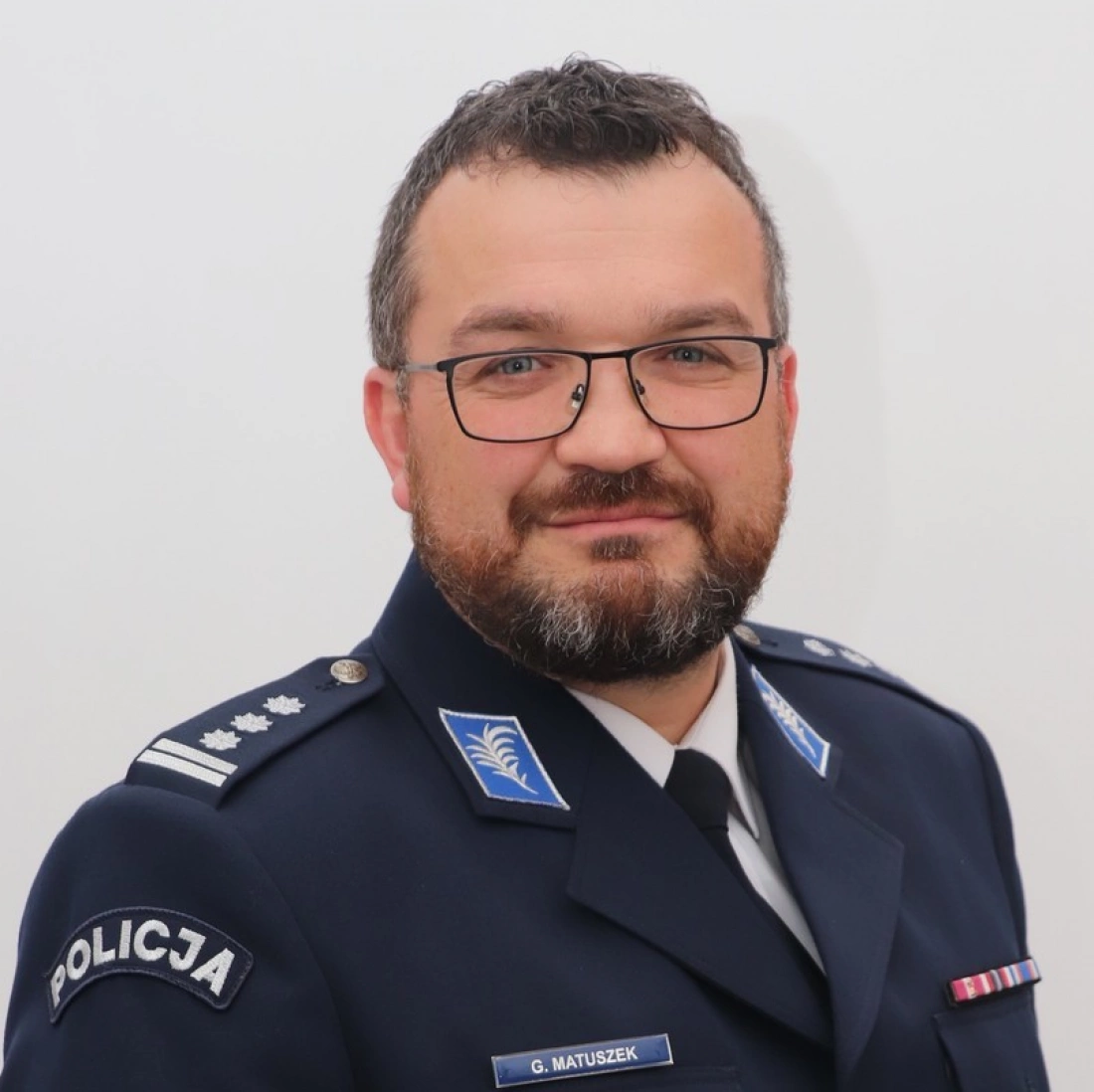 Mężczyzna w mundurze policyjnym z brodą, okularach, uśmiechający się.