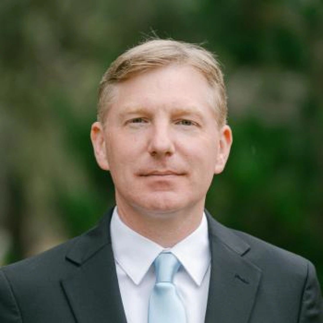 Mężczyzna w garniturze z jasnym krawatem na tle zieleni.