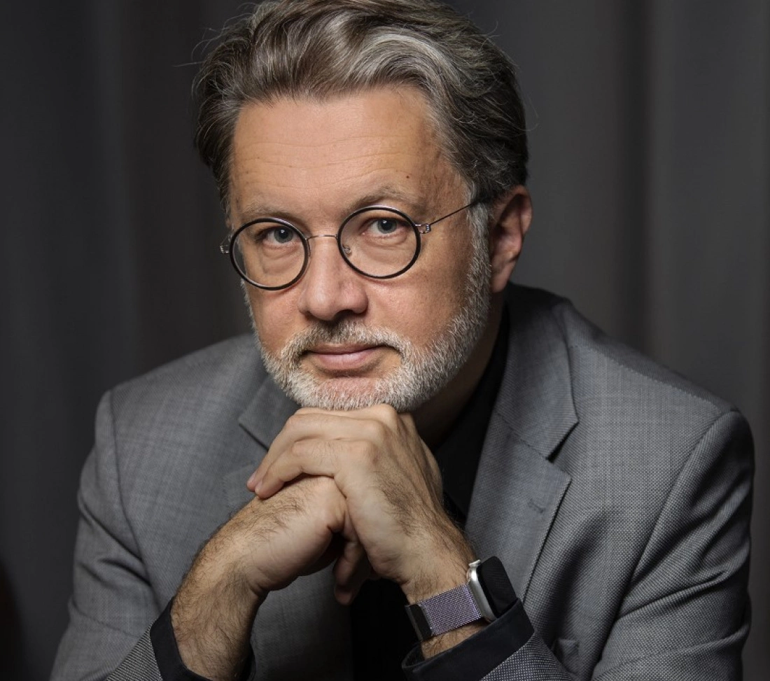 Mężczyzna w szarym garniturze i okrągłych okularach, z rękami złożonymi pod brodą, o spokojnym wyrazie twarzy.