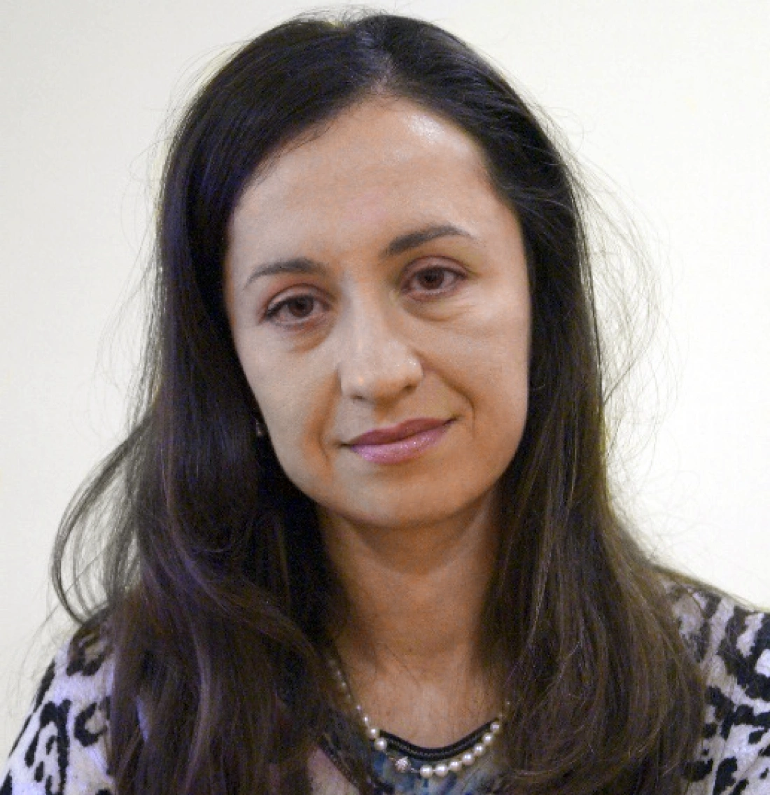 Kobieta o długich włosach, z delikatnym makijażem, uśmiecha się. W tle jasna ściana.