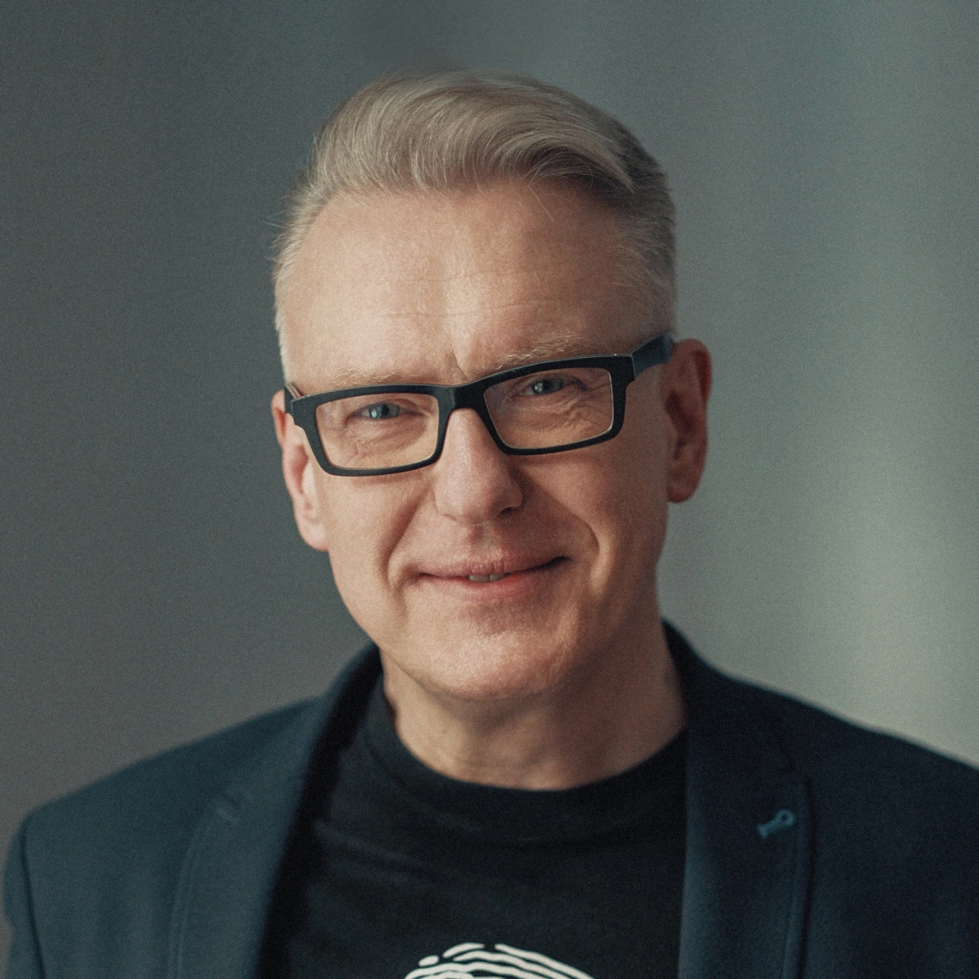 Mężczyzna w okularach, uśmiechający się, ubrany w czarną koszulkę i niebieską marynarkę.