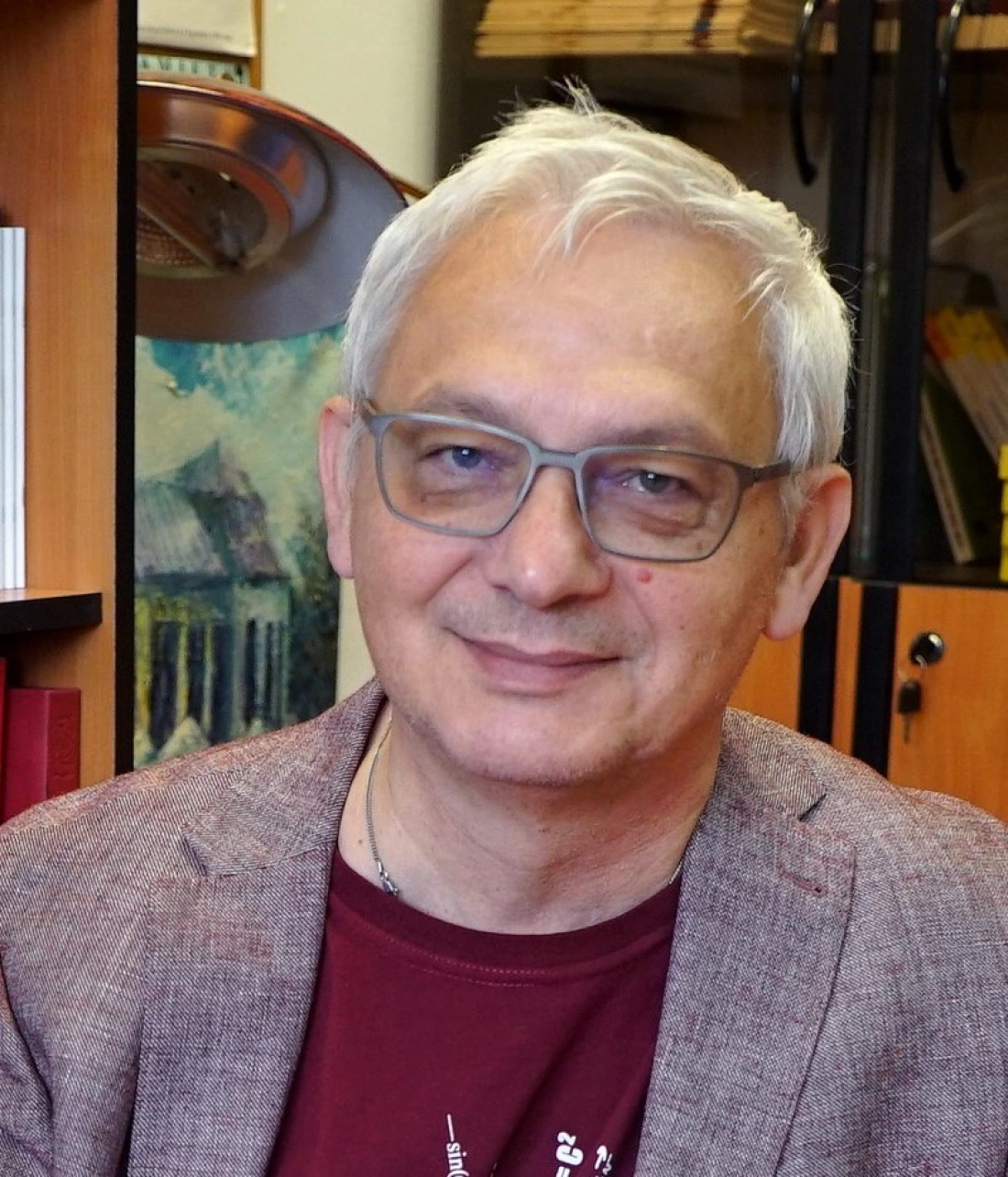 Mężczyzna z siwymi włosami i okularami, uśmiechający się, w otoczeniu regałów z książkami.