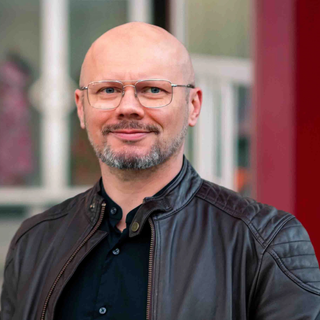 Mężczyzna z okularami, śmiejący się w ciężkiej kurtce, na tle nieostrego wnętrza.