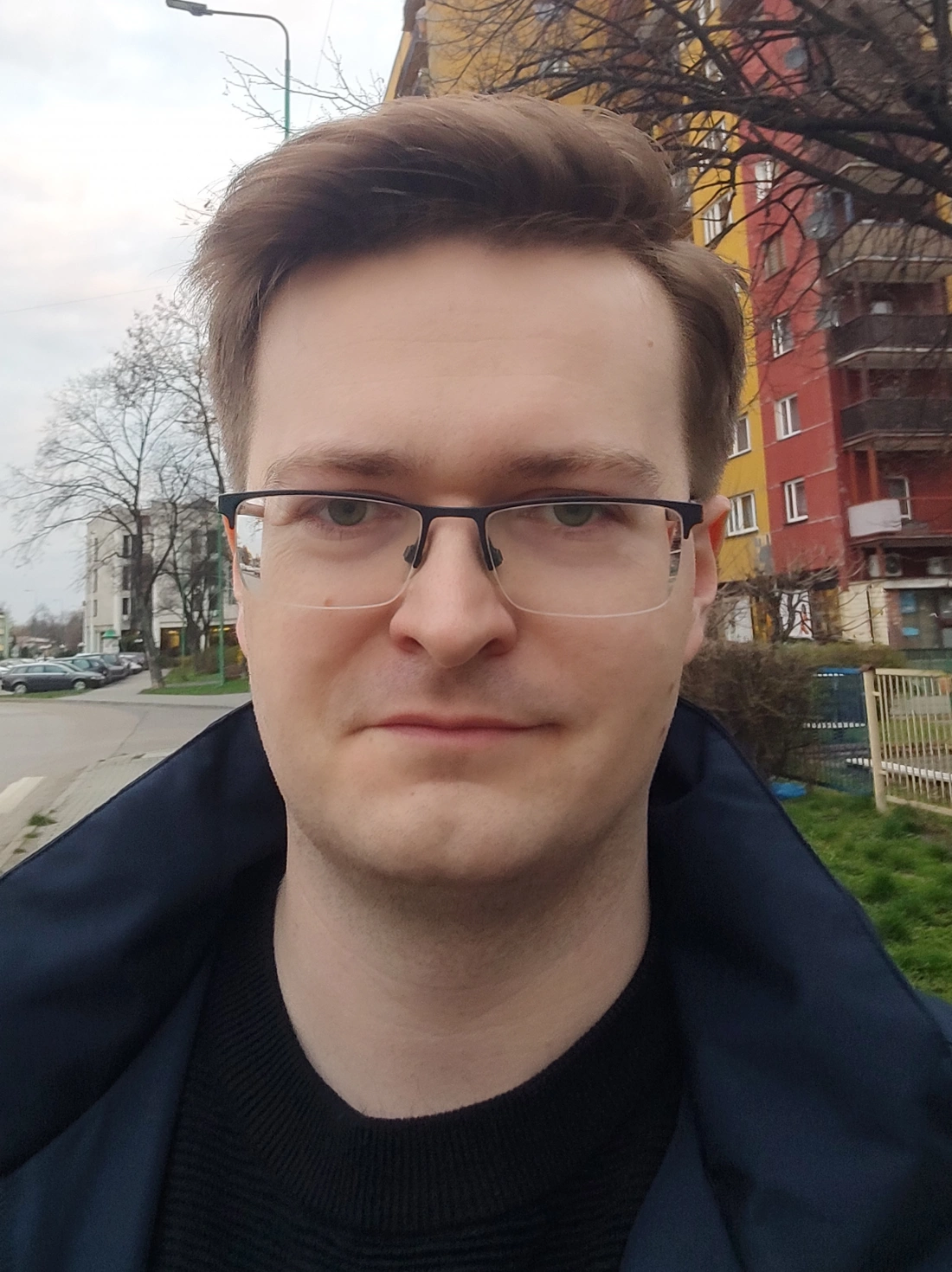 Mężczyzna o okularach stoi na ulicy, w tle kolorowe budynki.