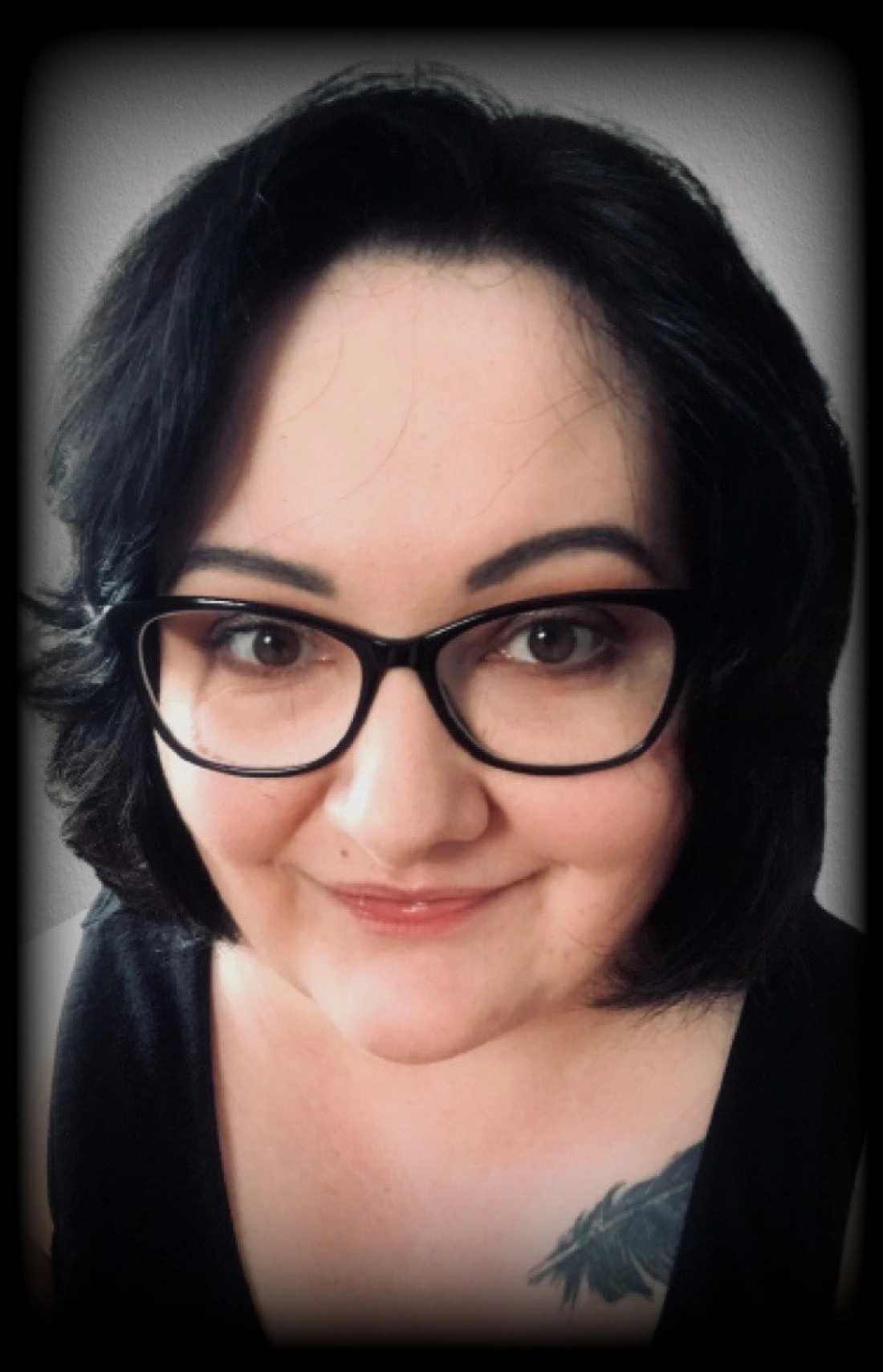 Portret kobiety w okularach, z krótkimi ciemnymi włosami i uśmiechem, z widocznym tatuażem na ramieniu.